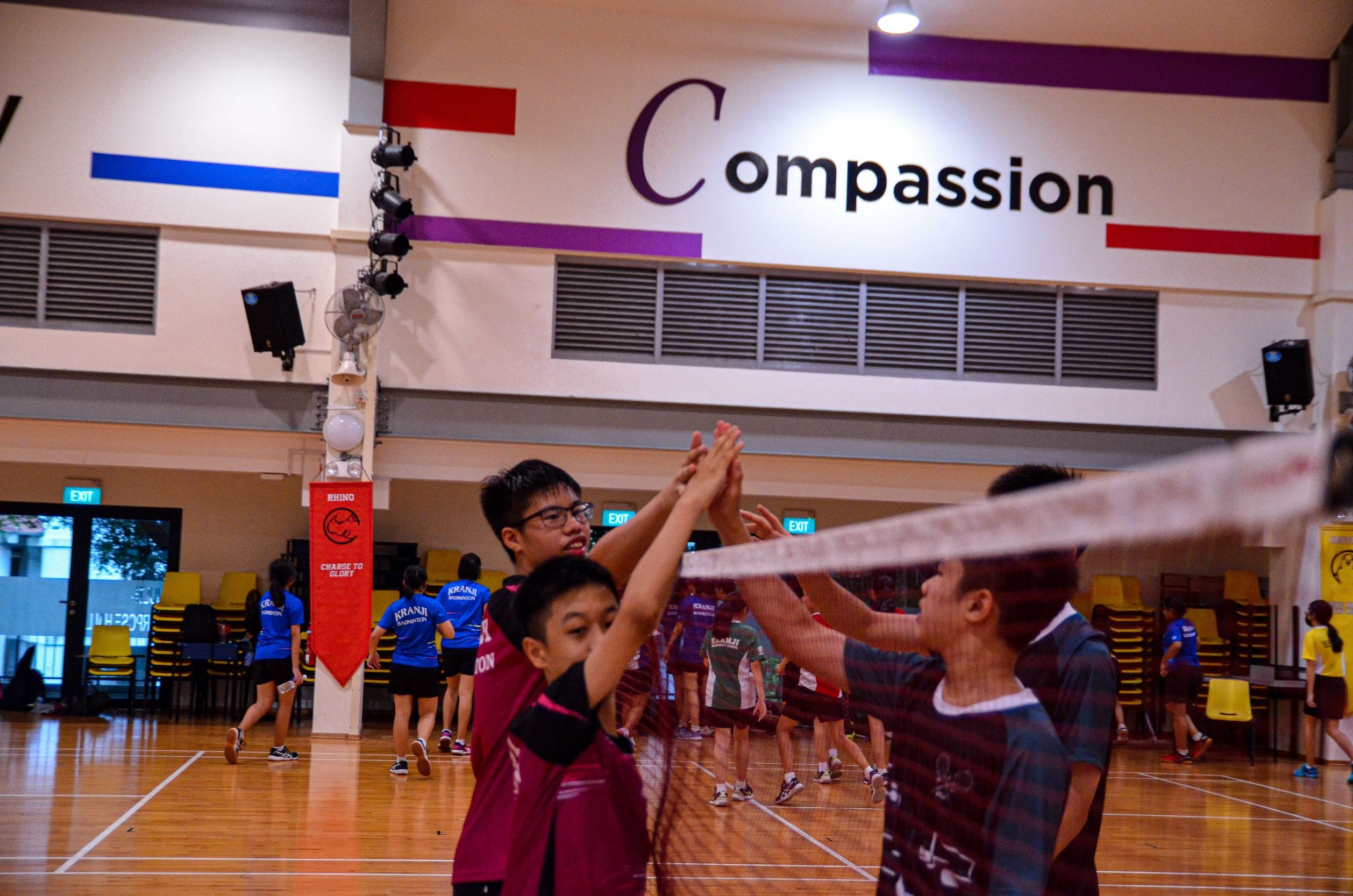 Youth players high-five over a badminton net indoors, with a wall sign reading Compassion in the background.